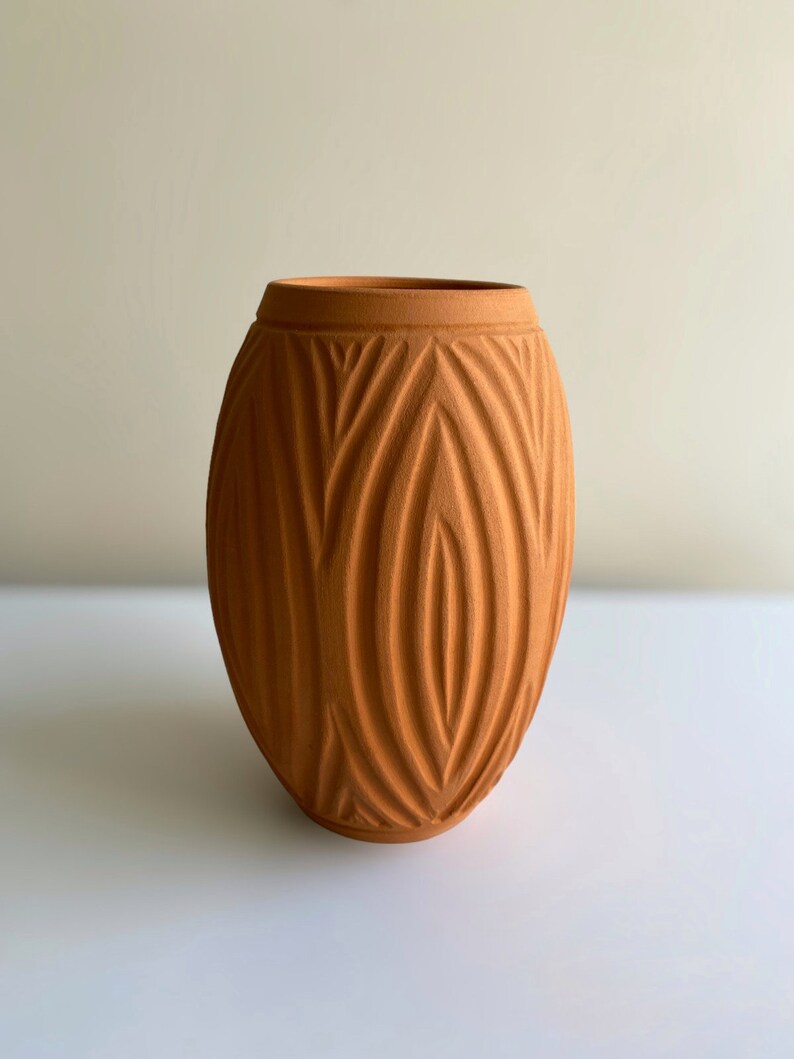 May include: A terracotta vase with a textured, geometric pattern. The vase is oval shaped and has a smooth, matte finish.