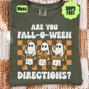 May include: A moss green t-shirt featuring a graphic of three white ghosts sitting at school desks in front of an orange and tan checkered background. The text reads ARE YOU FALL-O-WEEN DIRECTIONS? The shirt has a Comfort Colors brand label.