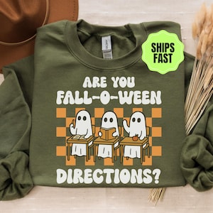 Könnte beinhalten: Ein olivgrünes Sweatshirt mit Rundhalsausschnitt und einer Grafik von drei weißen Geistern, die an Schulbänken sitzen. Der Text lautet ARE YOU FALL-O-WEEN DIRECTIONS? Das Design enthält ein orange-weißes Karomuster hinter den Geistern.