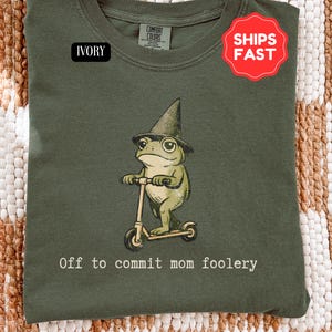 May include: An olive green t-shirt featuring a cartoon frog in a witch's hat on a scooter, with the text "Off to commit mom foolery". The shirt has a "Ships Fast" sticker. The t-shirt is labelled "Ivory".
