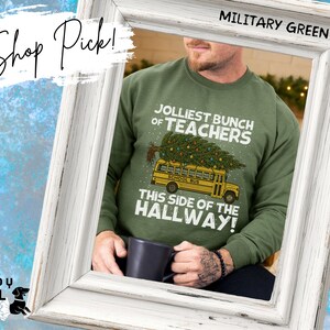 May include: A military green crewneck sweatshirt featuring a graphic of a yellow school bus carrying a decorated Christmas tree on its roof. The text reads JOLLIEST BUNCH OF TEACHERS THIS SIDE OF THE HALLWAY. A dark mug is held in the foreground.