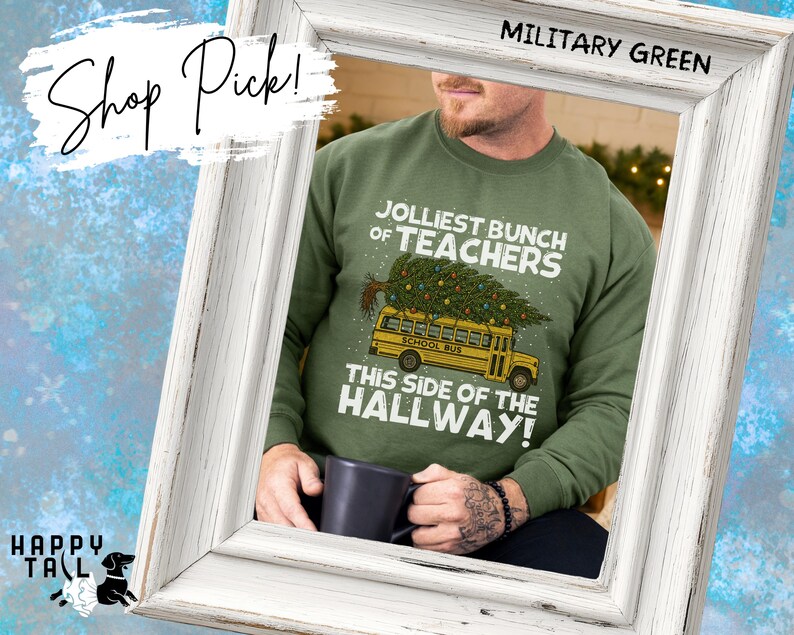 May include: A military green crewneck sweatshirt featuring a graphic of a yellow school bus carrying a decorated Christmas tree on its roof. The text reads JOLLIEST BUNCH OF TEACHERS THIS SIDE OF THE HALLWAY. A dark mug is held in the foreground.