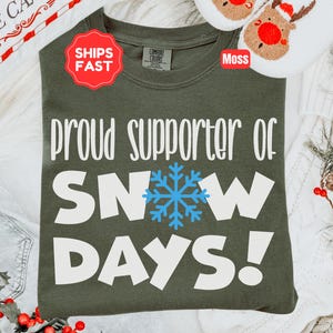 May include: A moss green crewneck t-shirt featuring the white text "PROUD SUPPORTER OF" above the white text "SNOW DAYS!" with a light blue snowflake graphic replacing the letter "O" in "SNOW". The shirt has a Comfort Colors brand label.