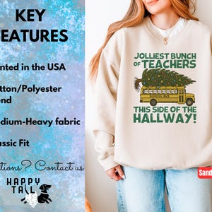 May include: A sand-colored crewneck sweatshirt featuring a graphic of a yellow school bus carrying a Christmas tree on its roof. The text reads JOLLIEST BUNCH OF TEACHERS THIS SIDE OF THE HALLWAY. The left side lists key features including printed in the USA.
