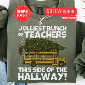 May include: A military green crewneck sweatshirt featuring a graphic of a yellow school bus carrying a decorated Christmas tree on its roof. The text reads JOLLIEST BUNCH OF TEACHERS THIS SIDE OF THE HALLWAY in white lettering with small white dots.