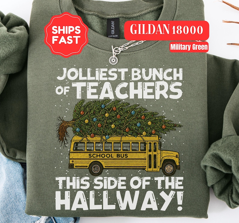 May include: A military green crewneck sweatshirt featuring a graphic of a yellow school bus carrying a decorated Christmas tree on its roof. The text reads JOLLIEST BUNCH OF TEACHERS THIS SIDE OF THE HALLWAY in white lettering with small white dots.