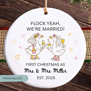 Könnte beinhalten: Runder weißer Weihnachtsbaumschmuck aus Keramik mit einem goldenen Faden. Der Schmuck zeigt zwei Cartoon-Gänse in Hochzeitskleidung, die Champagner halten. Der Text lautet "FLOCK YEAH, WE'RE MARRIED!" und "FIRST CHRISTMAS AS Mrs & Mrs Miller EST. 2025".