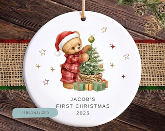 May include: A white ceramic Christmas ornament featuring a teddy bear in a red plaid outfit decorating a Christmas tree. The ornament has the text "Jacob's First Christmas 2025" and is decorated with stars and gifts. A personalized label is at the bottom.