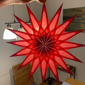 Puede incluir: Una gran estrella de papel roja con un diseño 3D. La estrella está colgada de una mano.