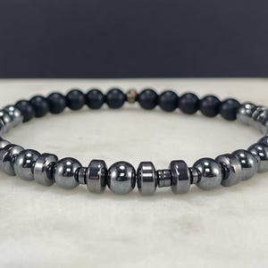 Könnte beinhalten: Ein Armband mit abwechselnd schwarzen und metallisch grauen Perlen. Das Armband besteht aus kleinen, runden Perlen und größeren, zylindrischen Perlen. Das Armband wird auf einer weißen Oberfläche präsentiert.