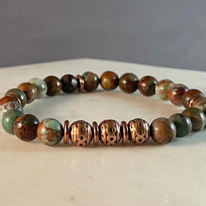 Pode incluir: Um bracelete feito com contas marrons e verdes com padrões e contas espaçadoras de cobre.