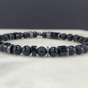 May include: Black beaded bracelet featuring a mix of matte and polished onyx beads, cylindrical and round, interspersed with silver-tone metal accents. The bracelet is strung on a durable cord and has a secure clasp.