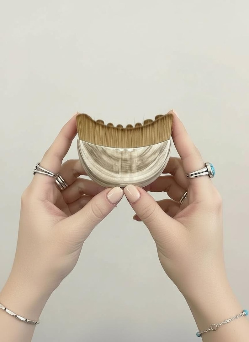 May include: A makeup brush with a curved, marbled handle and a fan-shaped brush head. The bristles are light brown. The handle is a light beige color with a marbled pattern. The brush is held between two hands.