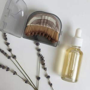 May include: A clear glass bottle with a white dropper, filled with a yellow liquid, sits next to a makeup brush in a gray case. Dried lavender sprigs are arranged on a white surface, creating a spa-like aesthetic.