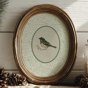 Puede incluir: Una impresión ovalada con marco dorado que presenta una ilustración de un pájaro verde. La impresión tiene un fondo verde claro con un patrón floral. El marco se coloca sobre una superficie de color neutro, con piñas y un tarro decorativo en primer plano.