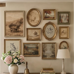 Op de afbeelding: Een galerijmuur met tien ingelijste kunstprints in verschillende vormen en maten, waaronder landschappen, een hert, een uil en een blad. Daaronder staat een houten sidetable met een witte vaas met roze bloemen, een koperen bel en boeken.
