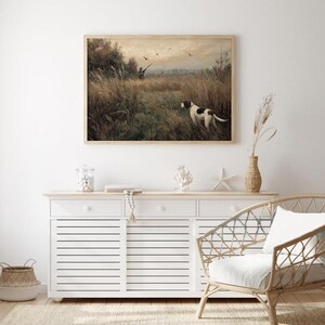 Peut inclure: Une impression vintage encadr&eacute;e d'une sc&egrave;ne de chasse avec un chien dans un champ, un chasseur et des oiseaux. L'&oelig;uvre est expos&eacute;e au-dessus d'un buffet blanc avec des objets d&eacute;coratifs et une chaise en osier.