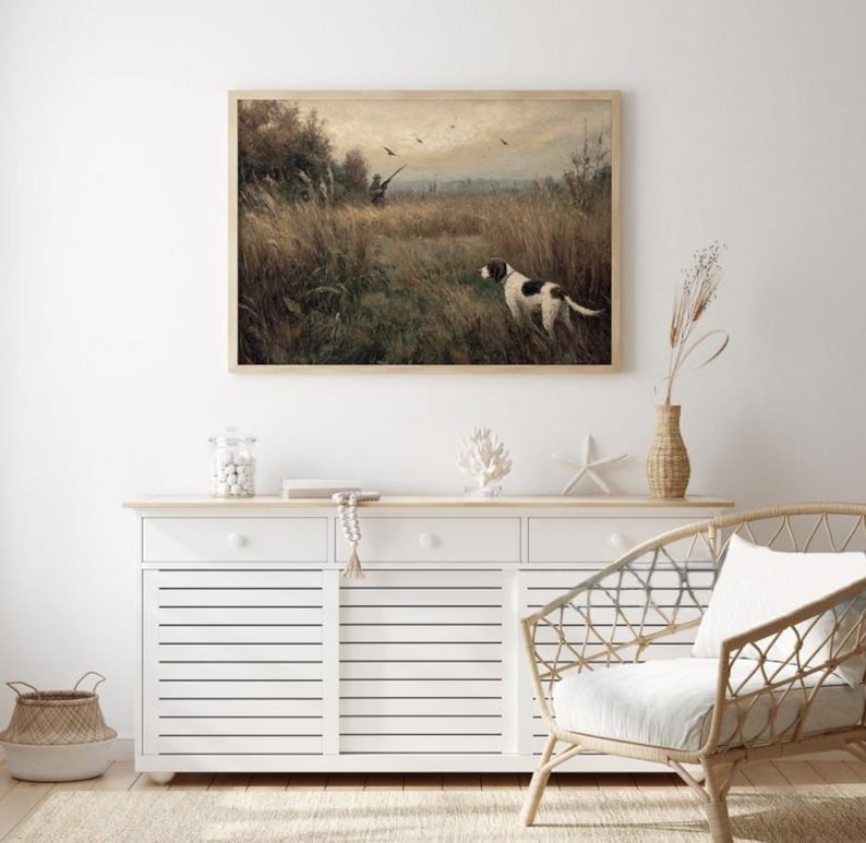 Peut inclure: Une impression vintage encadr&eacute;e d'une sc&egrave;ne de chasse avec un chien dans un champ, un chasseur et des oiseaux. L'&oelig;uvre est expos&eacute;e au-dessus d'un buffet blanc avec des objets d&eacute;coratifs et une chaise en osier.
