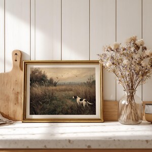 Peut inclure: Une impression vintage encadr&eacute;e repr&eacute;sentant une sc&egrave;ne de chasse avec un chien dans un champ. L'&oelig;uvre d'art est encadr&eacute;e d'or et est expos&eacute;e sur un comptoir avec une planche &agrave; d&eacute;couper en bois et un vase de fleurs s&eacute;ch&eacute;es. La palette de couleurs comprend des verts, des bruns et de l'or.