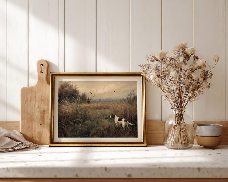 Peut inclure: Une impression vintage encadr&eacute;e repr&eacute;sentant une sc&egrave;ne de chasse avec un chien dans un champ. L'&oelig;uvre d'art est encadr&eacute;e d'or et est expos&eacute;e sur un comptoir avec une planche &agrave; d&eacute;couper en bois et un vase de fleurs s&eacute;ch&eacute;es. La palette de couleurs comprend des verts, des bruns et de l'or.