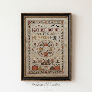 Op de afbeelding: Een ingelijste kruissteek sampler op lichte stof met de tekst GATHER ROUND, IT'S PUMPKIN HOUR. Het ontwerp bevat een centrale krans van pompoenen, eikels en bladeren, met een schuur, kaarsen en vleermuizen in een decoratieve rand.