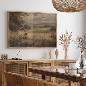 May include: A framed vintage print depicting a hunting scene with a dog in a field. The artwork features a dog, a hunter, and birds in flight, rendered in muted earth tones. The frame is made of wood.