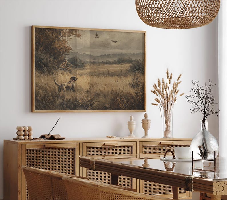 May include: A framed vintage print depicting a hunting scene with a dog in a field. The artwork features a dog, a hunter, and birds in flight, rendered in muted earth tones. The frame is made of wood.