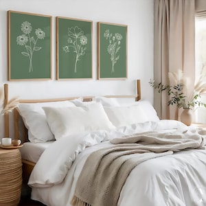 May include: Three framed prints featuring white line drawings of flowers on a sage green background. The prints are hanging on a white wall above a white bed with a beige throw blanket.