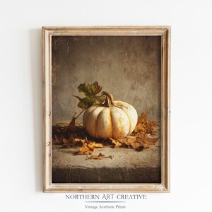 Op de afbeelding: Een ingelijste vintage print met een witte pompoen met oranje strepen, rustend op een bed van dorre bladeren. Een groen blad en een getextureerde achtergrond maken het herfststilleven compleet. De lijst is lichtbruin. De tekst "NORTHERN ART CREATIVE" en "Vintage Aesthetic Prints" staat onderaan.