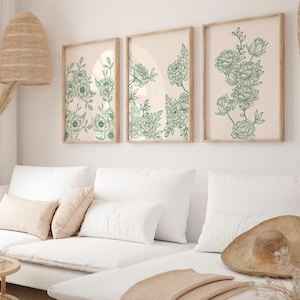 May include: Three framed prints with a white background and green floral illustrations. The prints are hanging on a white wall above a white sofa with cushions.