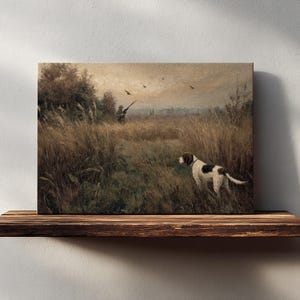 Peut inclure: Impression sur toile d'une sc&egrave;ne de chasse avec un chien dans un champ. Le chien, blanc avec des taches marron, se tient dans les hautes herbes. Un chasseur est visible &agrave; l'arri&egrave;re-plan, visant avec un fusil. Esth&eacute;tique vintage et rustique.