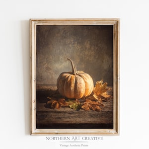 Puede incluir: Una impresión enmarcada que presenta una naturaleza muerta de una calabaza y hojas de otoño. La calabaza naranja está sobre una superficie de madera con hojas secas. La impresión tiene una estética vintage, con el texto "NORTHERN ART CREATIVE" y "Vintage Aesthetic Prints".