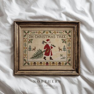 May include: A framed cross-stitch embroidery featuring Santa Claus carrying an axe and a pine tree, with the text OH CHRISTMAS TREE. The design includes holly borders, small deer, and houses in a muted colour palette, encased in a dark wood frame.