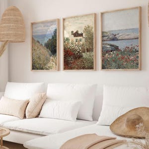 Puede incluir: Tres pinturas de paisajes enmarcadas con un sof&aacute; blanco y un sombrero de paja en primer plano. Las pinturas representan una casa en un campo de flores, un paisaje mar&iacute;timo con rocas y un camino a trav&eacute;s de un campo de flores.