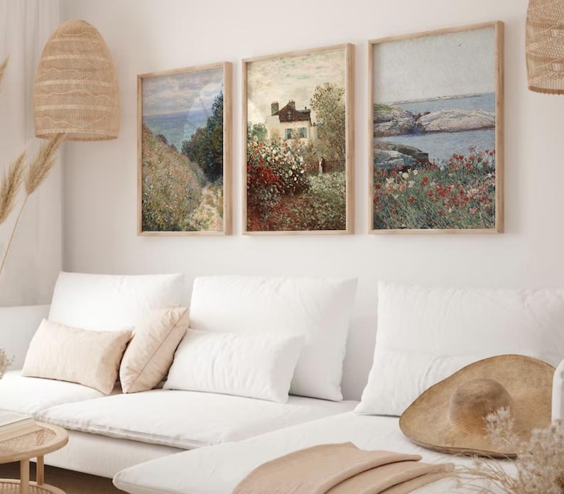 Puede incluir: Tres pinturas de paisajes enmarcadas con un sof&aacute; blanco y un sombrero de paja en primer plano. Las pinturas representan una casa en un campo de flores, un paisaje mar&iacute;timo con rocas y un camino a trav&eacute;s de un campo de flores.