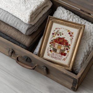 May include: A gold-toned rectangular frame containing a cross-stitch pattern of a basket filled with red apples, surrounded by colorful autumn leaves and a small owl, with the text AUTUMN HARVEST printed in a decorative red font at the bottom.