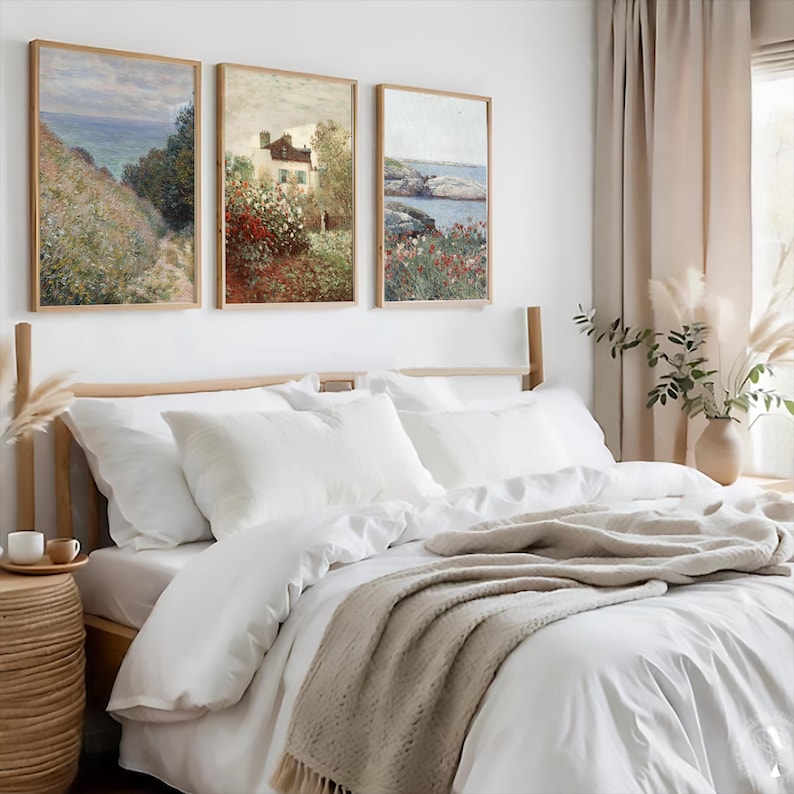 Puede incluir: Tres cuadros de paisajes enmarcados que representan paisajes marinos y una casa con jard&iacute;n, colgados en una pared blanca sobre una cama blanca con una manta beige y almohadas blancas.