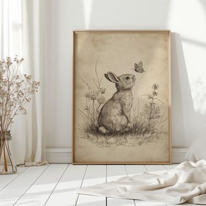 May include: A framed sepia-toned print of a rabbit gazing at a butterfly. The rabbit is sketched in detail, surrounded by grass and wildflowers. The artwork is displayed in a light-colored wooden frame, leaning against a white wall.