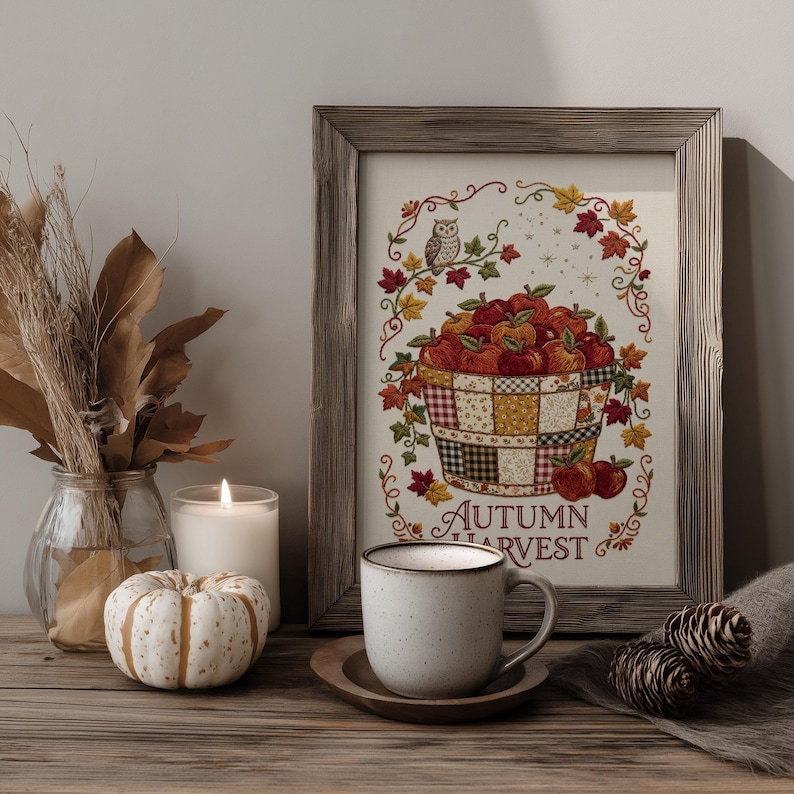 May include: A framed print featuring a basket of red apples, an owl, and autumn leaves with the text "AUTUMN HARVEST" in a decorative border. The artwork is displayed alongside a white speckled mug, a small white pumpkin, and dried foliage.