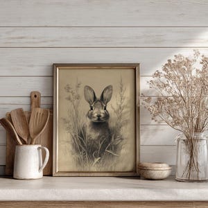 May include: A framed pencil sketch of a rabbit in a field of tall grass. The artwork is in a gold-colored frame and is displayed on a light-colored surface. A white ceramic mug with wooden utensils and a glass vase with dried flowers are also on the surface.