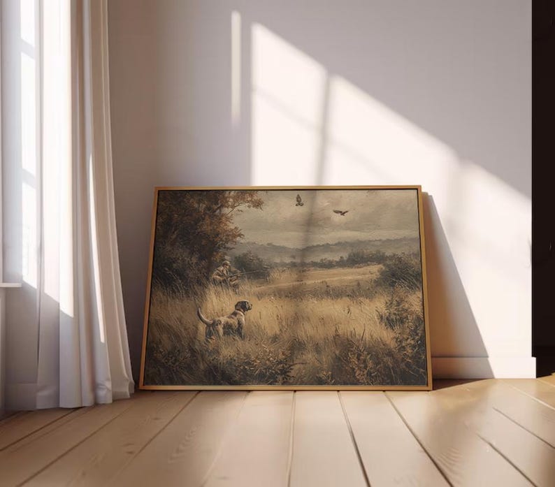 May include: A framed landscape painting featuring a hunting scene with a dog in a field of tall grass. The artwork has a muted color palette with a dog, hunter, and birds in the sky. The frame is a light brown color.