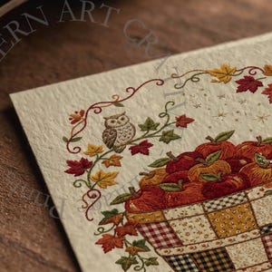 May include: A textured off-white card featuring an embroidered design of a woven basket filled with red and orange apples. An owl sits on a vine branch above the basket, surrounded by colorful autumn leaves and small gold star accents.