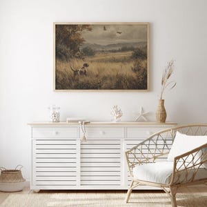May include: Framed landscape art print featuring a dog in a field, with trees and birds in the background. The artwork has a vintage aesthetic, with a neutral color palette and a wooden frame. The print is displayed above a white cabinet.