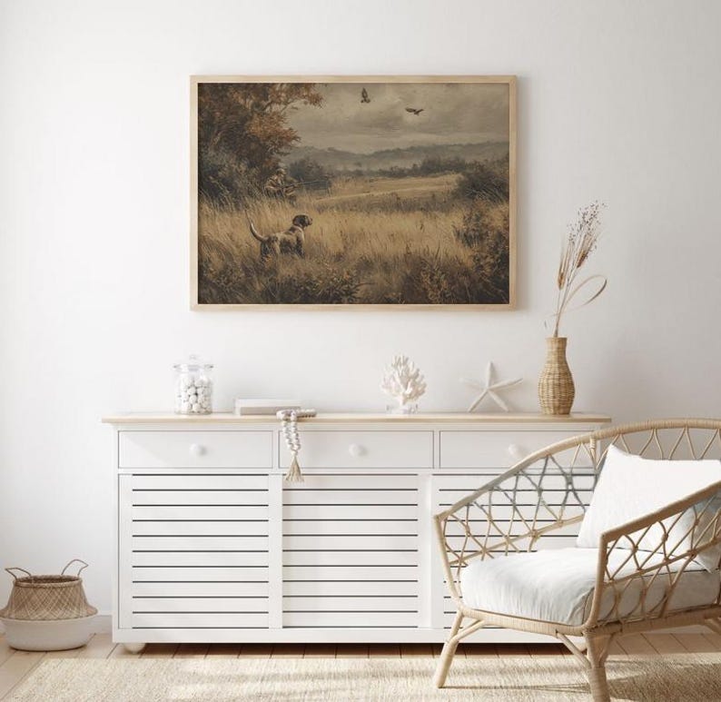 May include: Framed landscape art print featuring a dog in a field, with trees and birds in the background. The artwork has a vintage aesthetic, with a neutral color palette and a wooden frame. The print is displayed above a white cabinet.