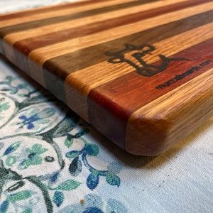 May include: A close-up of a wooden cutting board with a multi-colored striped design. The board has a rounded edge and is made of different types of wood. The board is branded with the text "mccardwood.com".