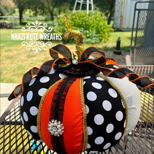 May include: A black and white polka dot pumpkin with orange and gold accents. The pumpkin has a black ribbon with a rhinestone embellishment and a gold stem.