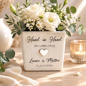 Könnte beinhalten: Ein Blumenarrangement in einem quadratischen, strukturierten, hellfarbigen Topf. Der Topf trägt die Worte "Hand in Hand" und ein Herzsymbol, darunter Namen und ein Datum. Die Blumen sind weiße Rosen und kleine weiße Blüten mit grünem Laub.