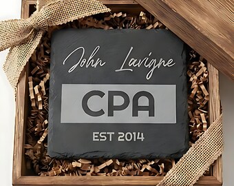 Op de afbeelding: Een gepersonaliseerde leistenen onderzetter met de gegraveerde naam "John Lavigne" en "CPA", plus "EST 2014". De onderzetter wordt gepresenteerd in een houten doos met een jute lint.