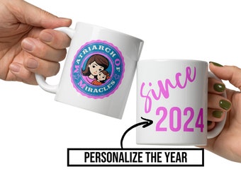 Op de afbeelding: Twee witte keramische mokken met roze en blauwe ontwerpen. Een mok heeft een blauwe cirkel met de tekst "Matriarch of Miracles" en een cartoonafbeelding van een moeder en kind. De andere mok heeft de tekst "since 2024" in roze.