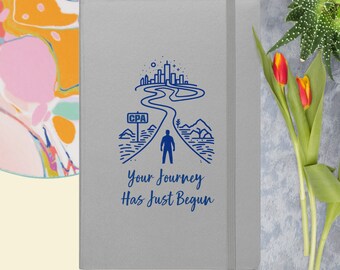 Op de afbeelding: Een grijs notitieboek met een blauwe illustratie van een persoon die naar een skyline loopt met de tekst "CPA" op een wegwijzer. De tekst "Your Journey Has Just Begun" staat onder de illustratie.
